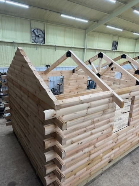 Log cabin under construction in workshop showing rounded pine logs and exposed roof trusses with metal brackets